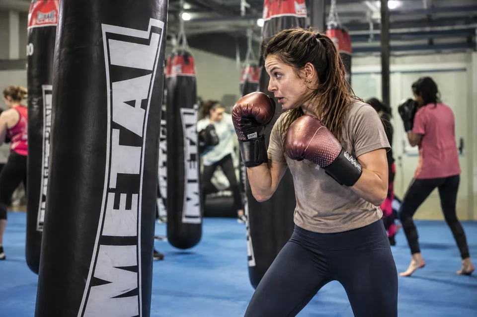 Boxing fitness près de Blagnac : une pratiquante en garde au sac, avec des gants bordeaux et une tenue taupe et marine.