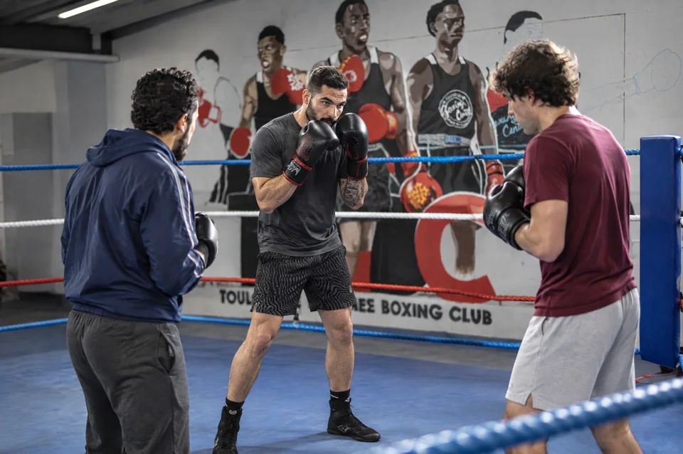 Salle de boxe proche de Blagnac : un coach montrant la garde à deux pratiquants sur un ring, devant la fresque des boxeurs.